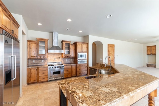 kitchen with built in appliances, light stone counters, wall chimney range hood, tasteful backsplash, and sink