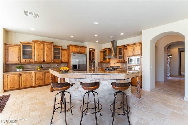 kitchen featuring a kitchen island with sink, a breakfast bar area, built in appliances, wall chimney exhaust hood, and decorative backsplash