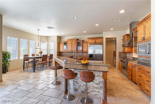 kitchen with a breakfast bar area, built in appliances, decorative backsplash, sink, and wall chimney range hood