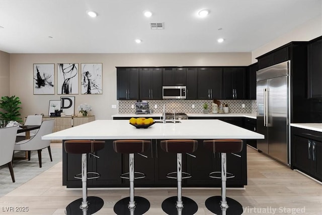 kitchen with a breakfast bar area, light hardwood / wood-style flooring, appliances with stainless steel finishes, an island with sink, and decorative backsplash