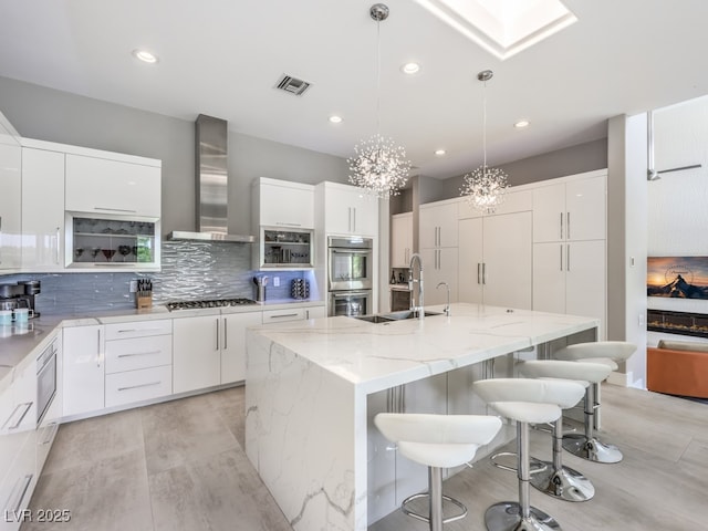 kitchen featuring appliances with stainless steel finishes, a kitchen island with sink, white cabinets, and wall chimney exhaust hood