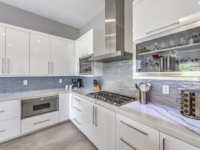 kitchen featuring backsplash, white cabinetry, stainless steel gas cooktop, light stone countertops, and wall chimney exhaust hood