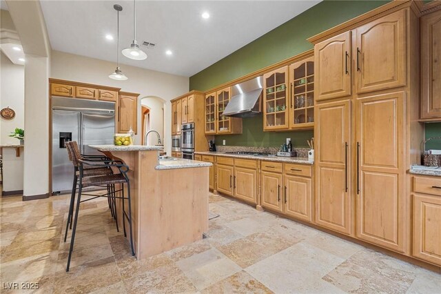 kitchen with stainless steel appliances, pendant lighting, light stone countertops, wall chimney range hood, and a kitchen island with sink