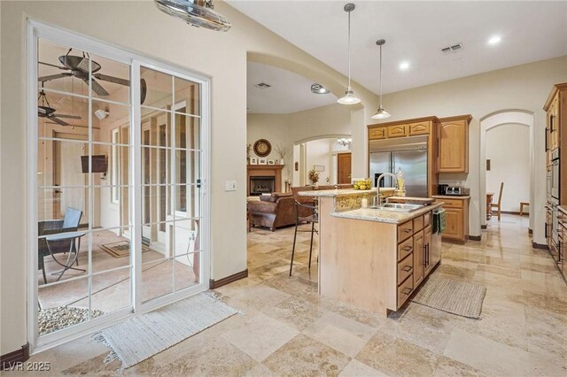 kitchen with a kitchen island with sink, appliances with stainless steel finishes, ceiling fan, light stone counters, and sink