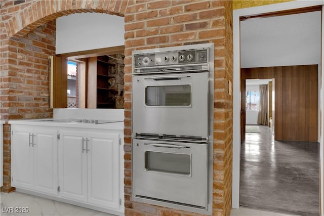 kitchen featuring brick wall, wooden walls, black electric stovetop, stainless steel double oven, and white cabinets