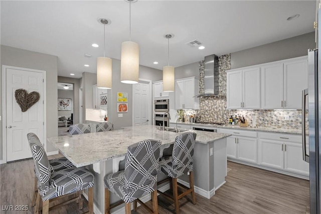 kitchen featuring hanging light fixtures, wall chimney range hood, a kitchen island with sink, white cabinetry, and a breakfast bar area