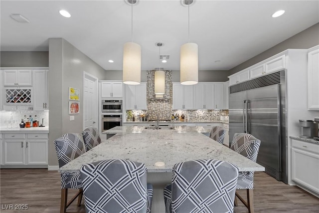 kitchen with a kitchen island with sink, stainless steel appliances, a breakfast bar, wall chimney range hood, and decorative light fixtures