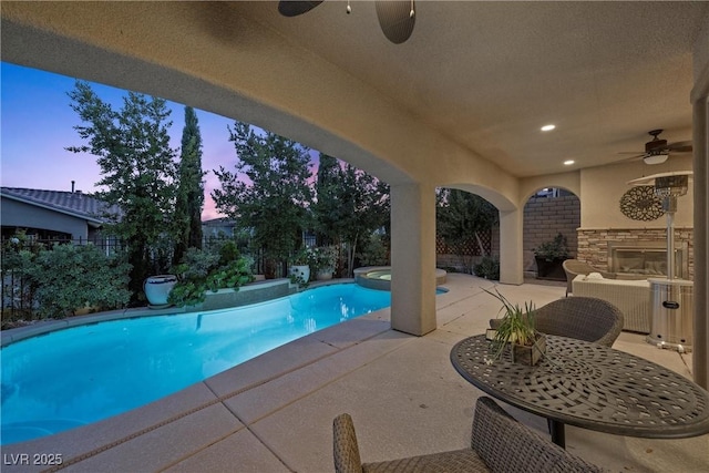 pool at dusk with a patio area, an outdoor stone fireplace, ceiling fan, and an in ground hot tub