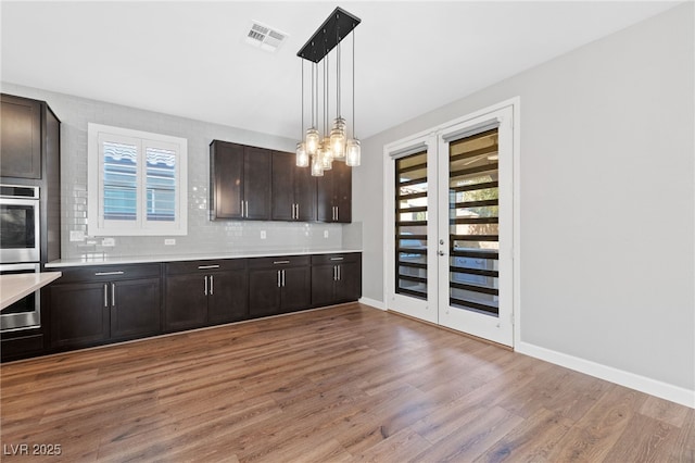 kitchen with wood-type flooring, hanging light fixtures, stainless steel double oven, dark brown cabinets, and backsplash