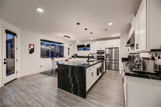 kitchen featuring decorative light fixtures, white cabinets, wall chimney exhaust hood, stainless steel fridge with ice dispenser, and a kitchen island with sink