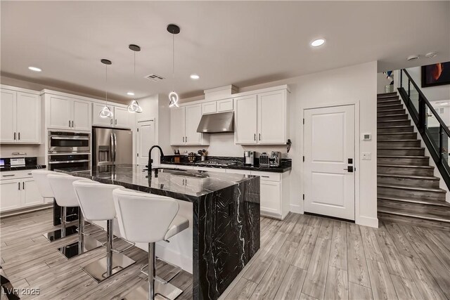 kitchen with a kitchen island with sink, appliances with stainless steel finishes, dark stone counters, light hardwood / wood-style flooring, and decorative light fixtures