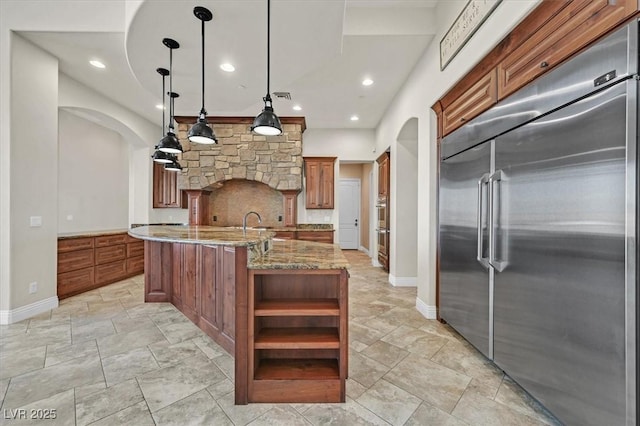 kitchen with light stone counters, open shelves, arched walkways, built in fridge, and brown cabinets