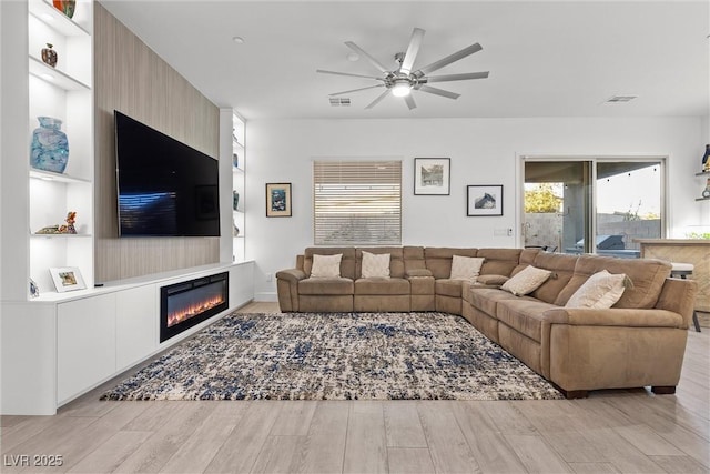 living room featuring ceiling fan, built in shelves, a large fireplace, and light hardwood / wood-style floors