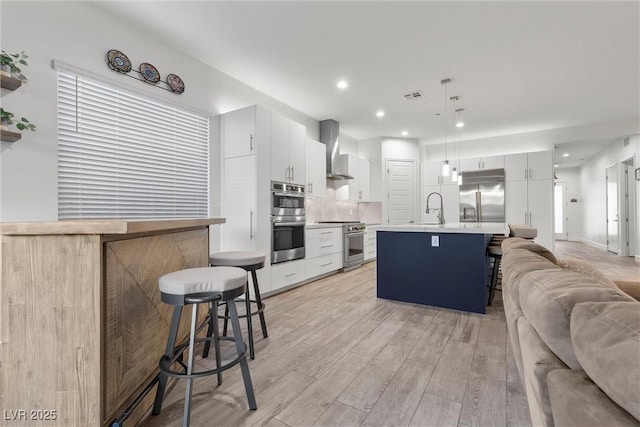 kitchen with premium appliances, a center island with sink, a breakfast bar area, pendant lighting, and wall chimney exhaust hood