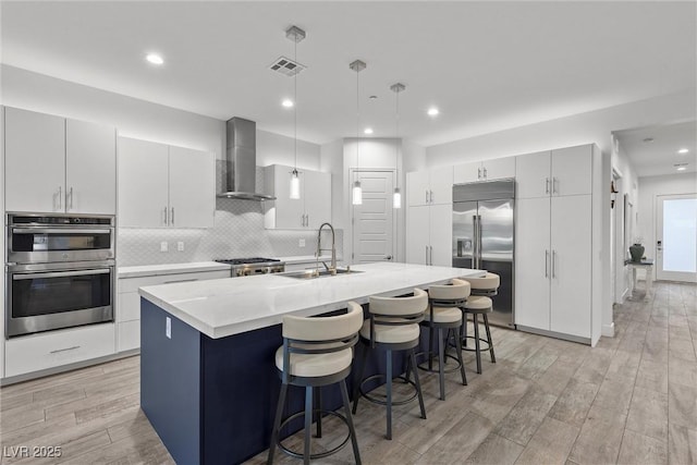 kitchen featuring wall chimney exhaust hood, stainless steel appliances, sink, hanging light fixtures, and a center island with sink