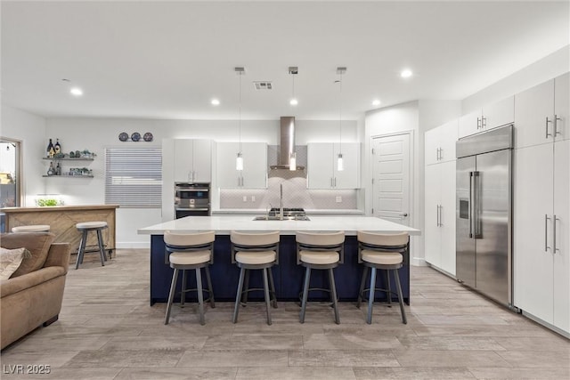 kitchen with decorative light fixtures, wall chimney range hood, a kitchen island with sink, appliances with stainless steel finishes, and white cabinets