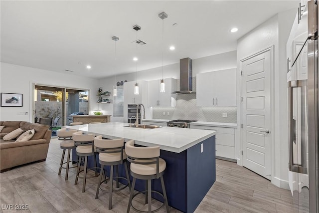 kitchen with a center island with sink, wall chimney range hood, pendant lighting, white cabinets, and sink