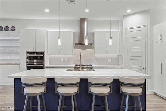 kitchen with white cabinetry, a center island with sink, stainless steel double oven, and wall chimney range hood