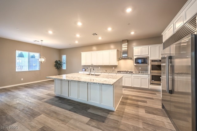 kitchen with a center island with sink, white cabinets, wall chimney range hood, and appliances with stainless steel finishes