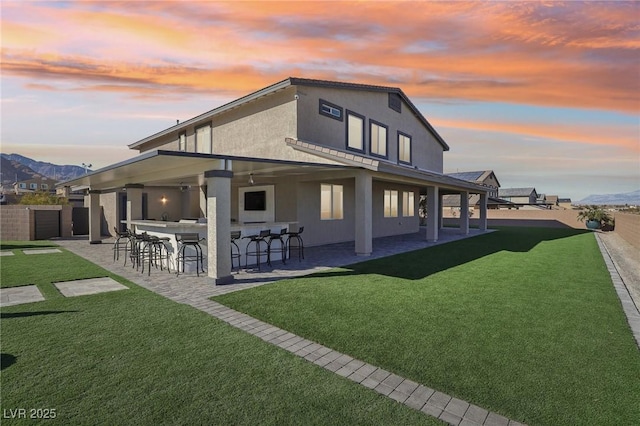 back house at dusk featuring a patio area, exterior bar, a lawn, and a mountain view