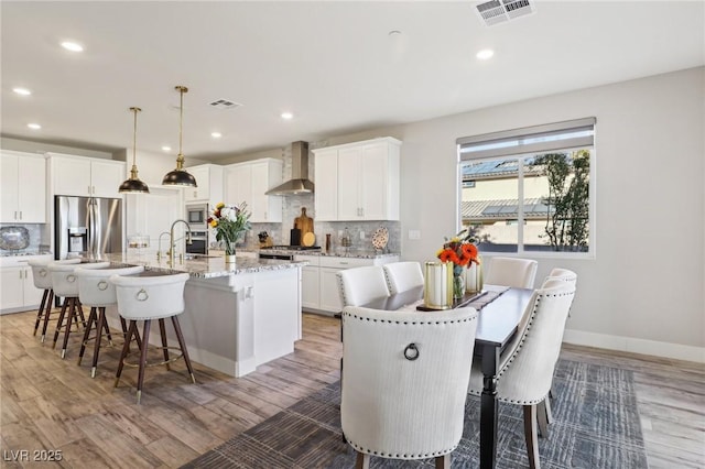 kitchen with white cabinetry, a kitchen island with sink, wall chimney range hood, pendant lighting, and stainless steel appliances