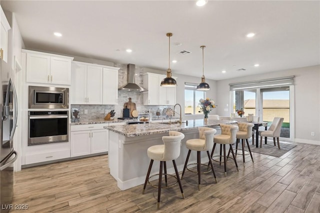 kitchen featuring pendant lighting, wall chimney range hood, appliances with stainless steel finishes, and an island with sink