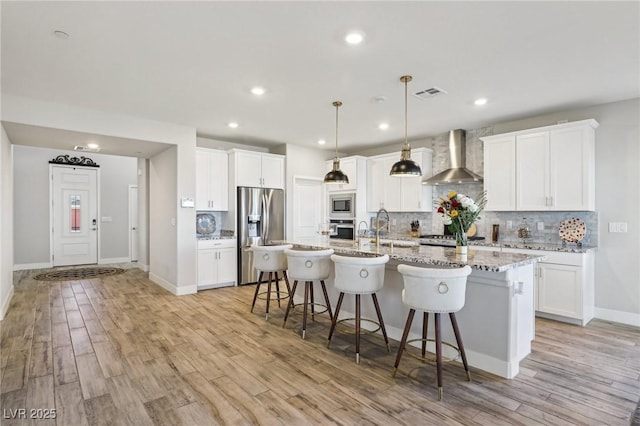 kitchen with white cabinetry, appliances with stainless steel finishes, wall chimney exhaust hood, and an island with sink