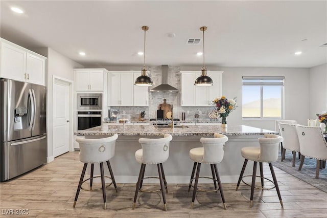 kitchen with wall chimney exhaust hood, decorative light fixtures, a center island with sink, and appliances with stainless steel finishes