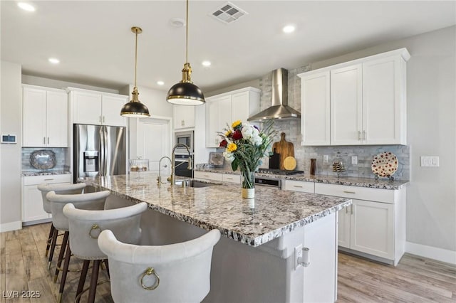 kitchen featuring white cabinetry, wall chimney range hood, pendant lighting, stainless steel appliances, and a center island with sink