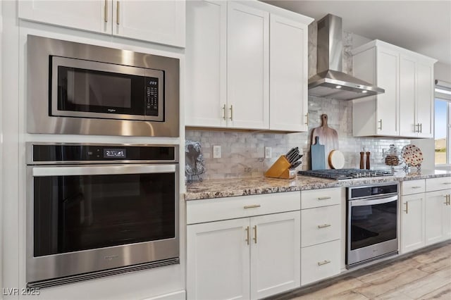 kitchen with decorative backsplash, wall chimney range hood, white cabinets, stainless steel appliances, and light stone counters