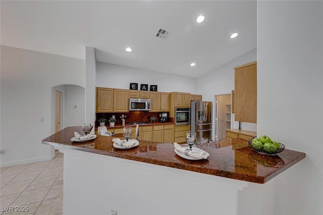 kitchen featuring light tile patterned flooring, kitchen peninsula, decorative backsplash, dark stone counters, and appliances with stainless steel finishes