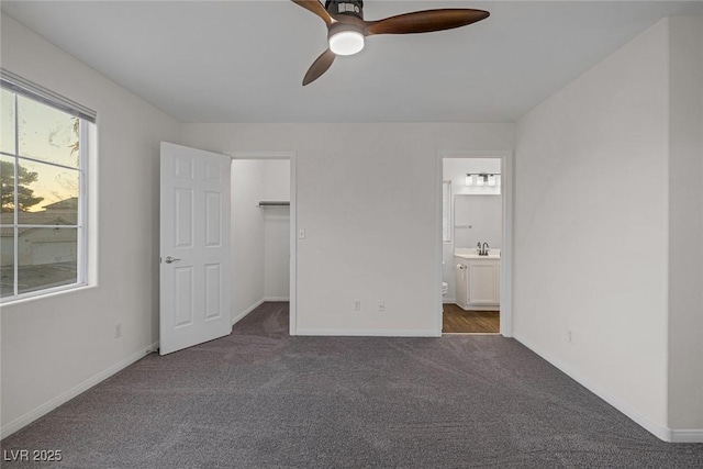 unfurnished bedroom with ensuite bath, a closet, a walk in closet, ceiling fan, and dark carpet