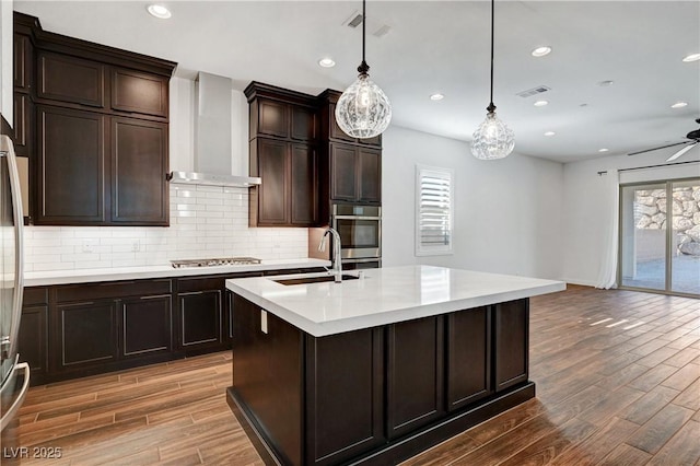 kitchen with wall chimney range hood, plenty of natural light, sink, hanging light fixtures, and a kitchen island with sink