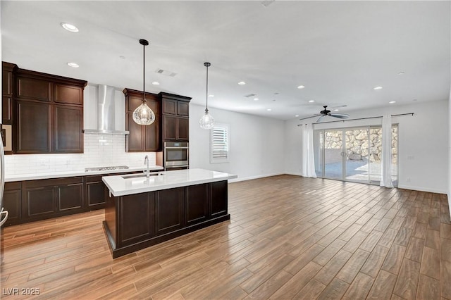 kitchen featuring ceiling fan, hanging light fixtures, a kitchen island with sink, stainless steel appliances, and wall chimney exhaust hood