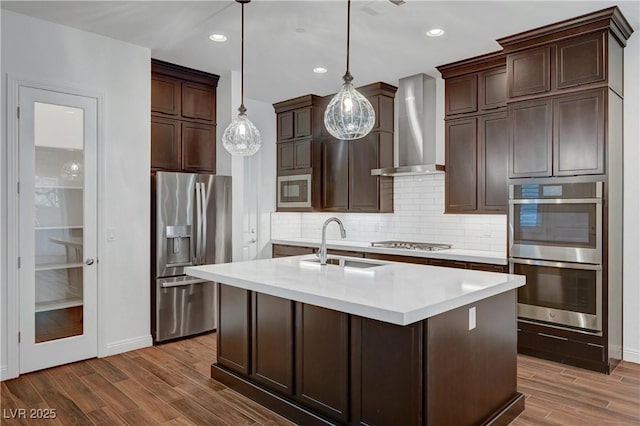 kitchen with a center island with sink, sink, hanging light fixtures, appliances with stainless steel finishes, and wall chimney exhaust hood