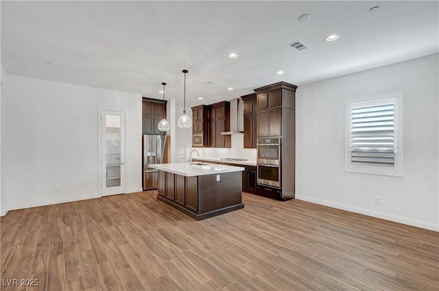 kitchen with pendant lighting, appliances with stainless steel finishes, wall chimney range hood, sink, and a center island with sink