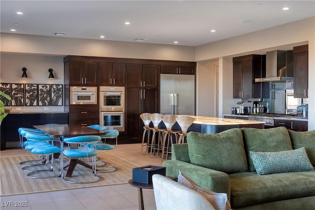 kitchen featuring stainless steel appliances, a kitchen bar, dark brown cabinets, wall chimney range hood, and light tile patterned flooring