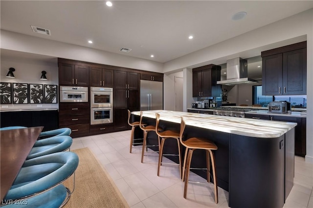 kitchen featuring stainless steel appliances, dark brown cabinetry, a kitchen island, wall chimney range hood, and a breakfast bar