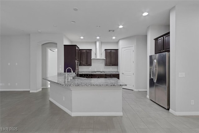 kitchen with a center island with sink, dark brown cabinetry, stainless steel fridge with ice dispenser, gas stovetop, and wall chimney exhaust hood