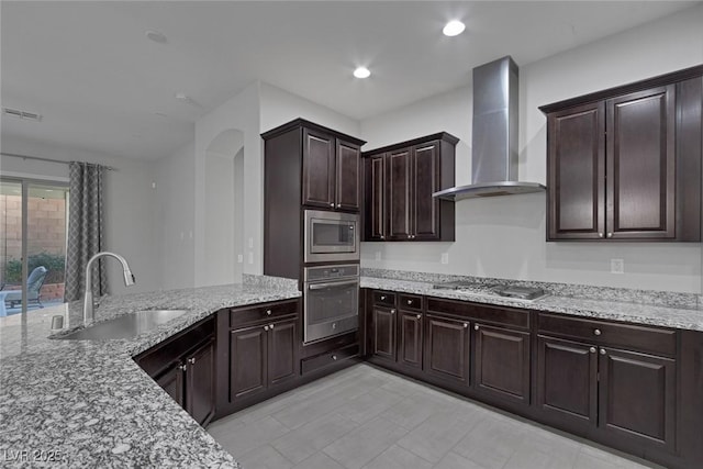 kitchen featuring light stone countertops, appliances with stainless steel finishes, wall chimney exhaust hood, sink, and dark brown cabinets