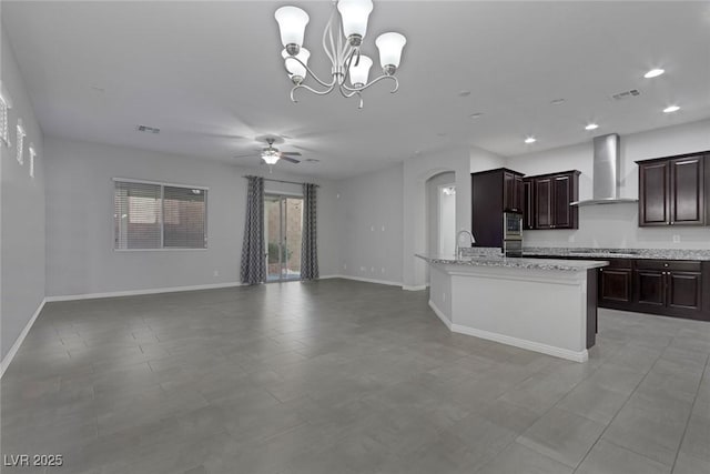 kitchen featuring pendant lighting, dark brown cabinets, light stone countertops, wall chimney exhaust hood, and ceiling fan with notable chandelier