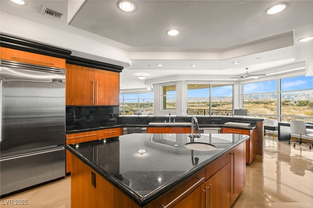 kitchen featuring an island with sink, ceiling fan, stainless steel appliances, dark stone counters, and sink