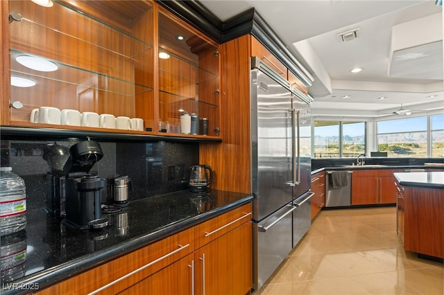 kitchen with light tile patterned floors, stainless steel appliances, a tray ceiling, dark stone countertops, and sink