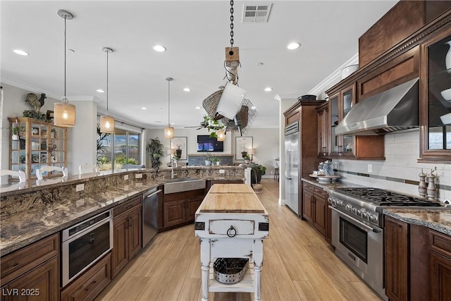 kitchen with butcher block counters, premium appliances, decorative light fixtures, wall chimney range hood, and sink