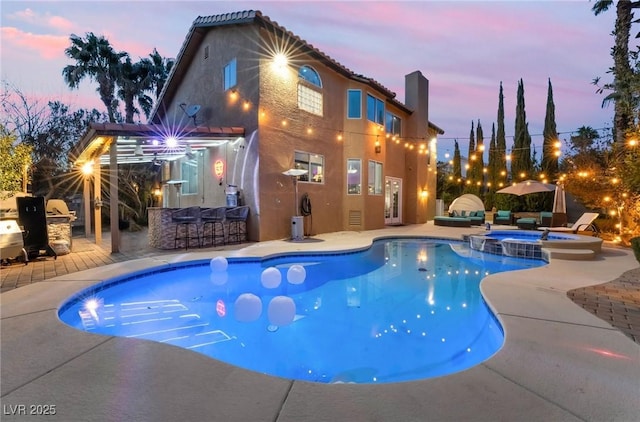 pool at dusk featuring an outdoor kitchen, a bar, an in ground hot tub, and a patio