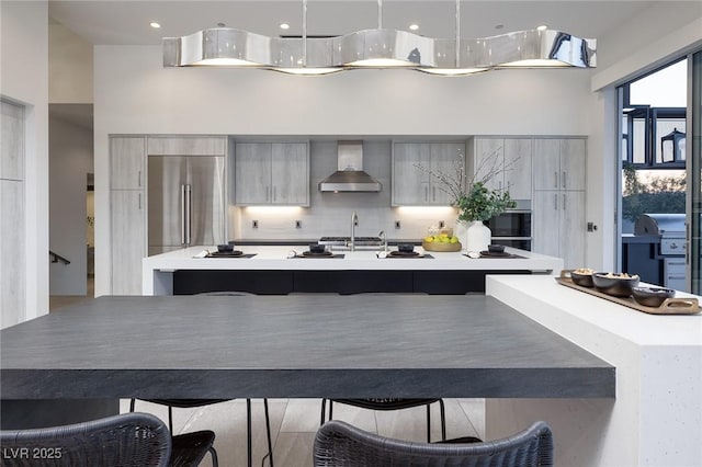 kitchen with built in refrigerator, pendant lighting, wall chimney exhaust hood, a kitchen bar, and sink