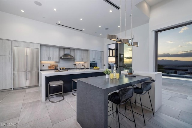 kitchen with an island with sink, a breakfast bar area, stainless steel appliances, pendant lighting, and wall chimney exhaust hood