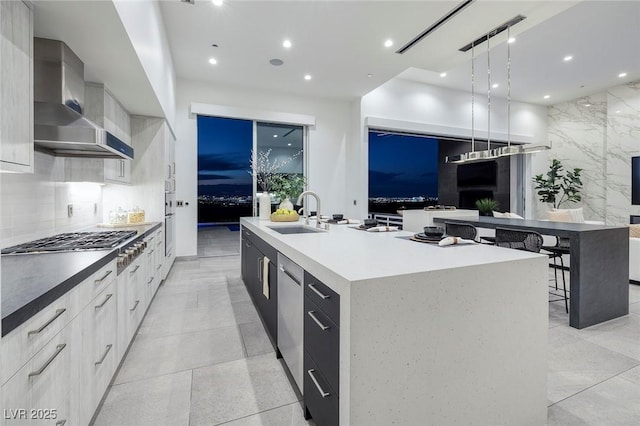 kitchen with a large island with sink, wall chimney exhaust hood, decorative light fixtures, sink, and stainless steel gas cooktop