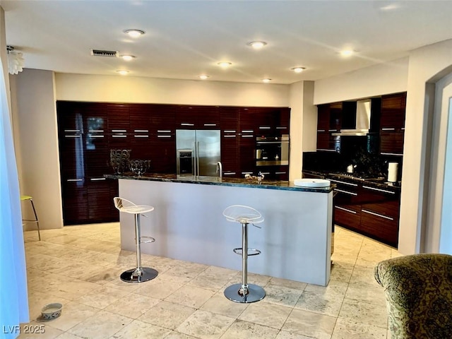 kitchen featuring a large island, a breakfast bar area, stainless steel appliances, dark stone countertops, and wall chimney range hood