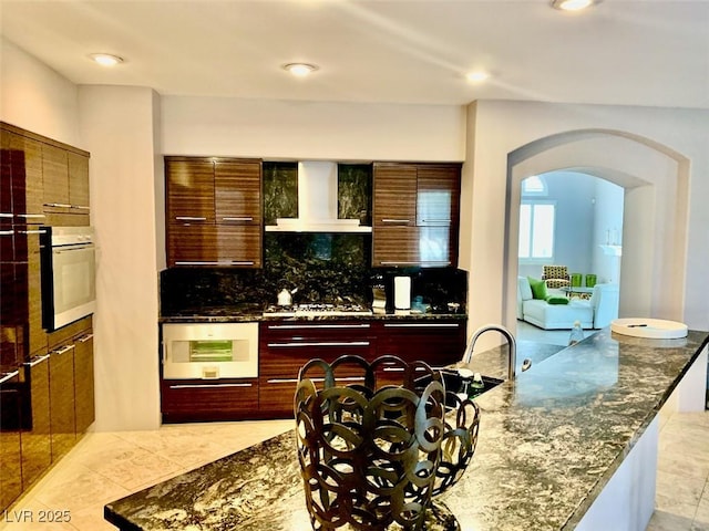 kitchen featuring backsplash, dark stone counters, stainless steel oven, white oven, and wall chimney exhaust hood